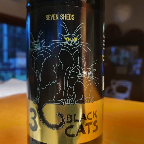 Image of a label on a beer bottle sitting on a bar. The label is black with Seven Sheds written in metallic gold. The label shows pale outline sketches of three quite smug-looking black cats, on a black background. One cat has green eyes, one has yellow eyes and one has blue eyes. A gold band around the bottom of the label is the background for the tail of one cat and on the gold band are the words 3 Black Cats.