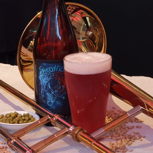 A bottle and a glass of Swordfish Trombones beer framed by a trombone, sitting on a cloth with a small dish of hop pellets and scattered barley.