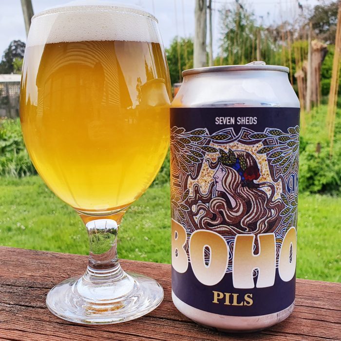 A thick-stemmed glass with beer in it, next to a can with a mosaic image of a woman with barley and hops, Seven Sheds Boho Pils.